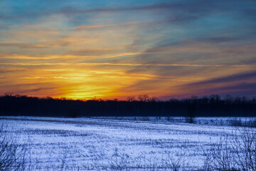 A bright sun disk disappears behind the horizon, painting the sky with rich gold, orange, and blue hues. The snowy field and dark forest line create a vivid contrast that emphasizes the grandeur of th © Олег Струс
