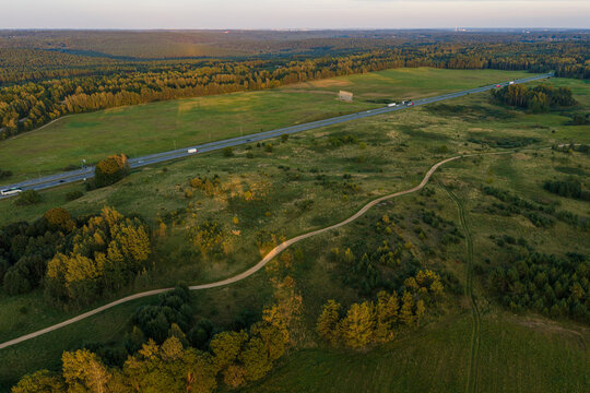 Aerial view of a winding dirt path through green fields and forest patches alongside a highway with vehicles in the golden hour light in Ziezmariai, Kaunas County, Lithuania.