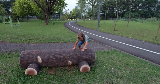 Adventurous toddler cautiously climbing onto onto faux wood concrete log bench and beam walking along it with arms outstretched, demonstrating purposeful movement and exploring textures barefoot