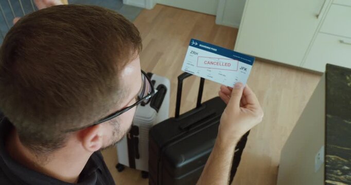 Frustrated traveler shaking his head while looking at a cancelled boarding pass with luggage, reacting to unexpected flight cancellation.