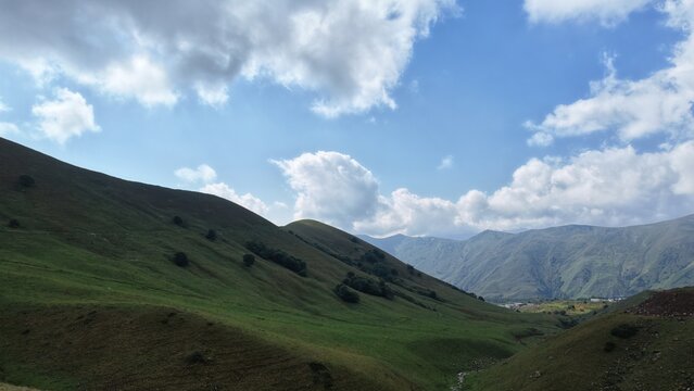 Wide panoramic landscape of a lush green valley nestled between rolling mountain slopes, with a small stream visible below and dramatic clouds overhead.