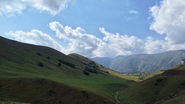 Sweeping panoramic view of smooth green rolling hills and valleys leading toward distant mountain ranges, with fluffy white clouds filling the sky above.
