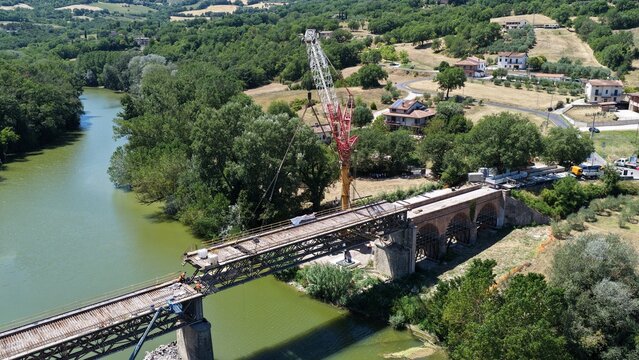 Aerial view of a bridge under construction with a large crane positioned over the river, surrounded by lush green trees and rural landscape in Todi, Umbria, Italy.