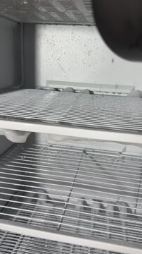 Homeowner defrosts freezer, melting thick ice with a hair dryer's warm airflow, water dripping into towels, candid authentic life chore at home in a quiet kitchen this afternoon.