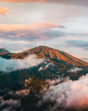 Aerial view of the lush mountainous landscape of East Java at golden hour, with soft clouds drifting through the forested slopes in Kecamatan Pujon, Jawa Timur, Indonesia.