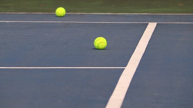 Two neon yellow tennis balls resting on a blue hard court with white boundary lines during a sports practice session