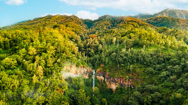 Aerial view of Coban Rondo Waterfall surrounded by lush green tropical forest and rocky cliffs in Kecamatan Pujon, Jawa Timur, Indonesia.