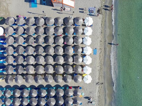 Aerial view of the beach in Katarinies Paralia, Greece, featuring rows of straw umbrellas and sun loungers on the sand next to the clear turquoise sea. Katarinies Paralia, Greece.
