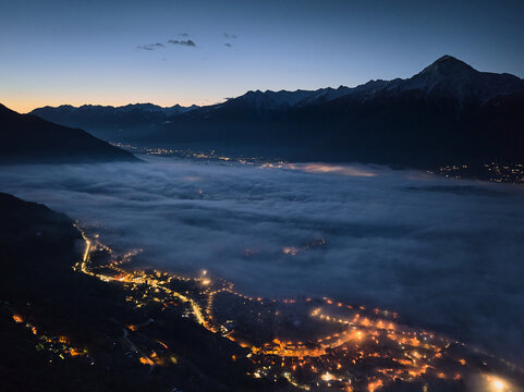 Aerial view of Valtellina valley at night with city lights glowing through thick fog, surrounded by dark mountain silhouettes under a clear twilight sky in Sondrio, Lombardy, Italy.