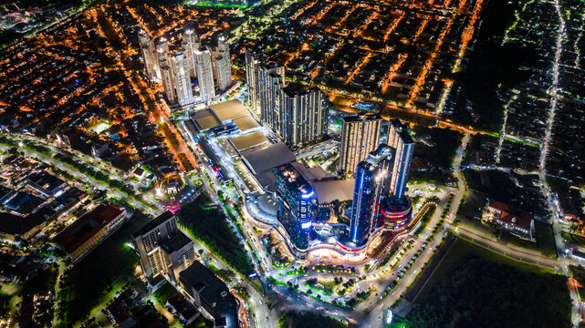 Aerial view of the illuminated cityscape of Surabaya at night, featuring high-rise apartment towers, a large shopping mall complex, and glowing street lights in Surabaya, East Java, Indonesia.