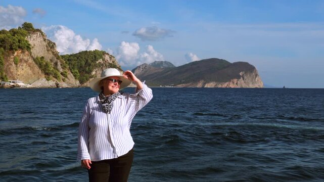 Mature Woman Enjoying Seaside Vacation by the Mountains. Mature woman in sun hat and sunglasses standing by the sea and enjoying a sunny coastal vacation with mountains in the background.