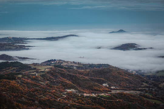 Aerial view of the village of Sarnano nestled among rolling hills covered in autumn foliage and partially obscured by a thick sea of clouds, Sarnano, Marche, Italy.