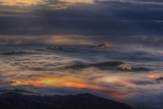 Aerial view of the town of Sarnano illuminated by warm lights piercing through a thick blanket of fog covering the rolling hills of the Marche region at night Sarnano, Marche, Italy.