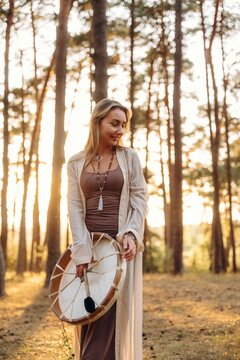 Young woman in brown dress holds traditional drum while standing in sunlit forest, surrounded by tall trees and golden grass, creating a serene outdoor atmosphere