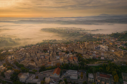 Aerial view of Corridonia with its historic rooftops and church towers emerging from the morning fog at sunrise in the rolling hills of the Marche region, Corridonia, Marche, Italy.