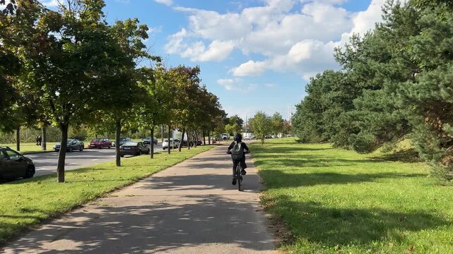 Child riding bicycle on paved path on city street under blue sky with scattered clouds, surrounded by green grass and trees. Sunlit outdoor scene with child cycling away from camera on tree-lined path