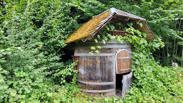 Small rustic wooden hut designed in the shape of a wine barrel, surrounded by lush green vegetation in the Swiss countryside. The unusual wooden structure and natural setting create a charming and aut