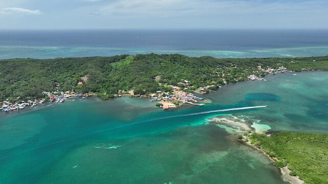 Aerial view of Isla Santa Elena featuring lush green hills, clear turquoise waters, a coastal village, and a boat leaving a wake in Jose Santos Guardiola, Islas de la Bahia, Honduras.