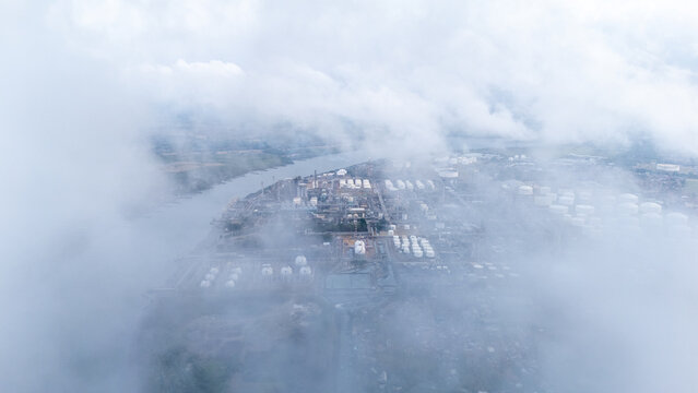 Aerial view of an industrial refinery complex situated along a riverbank, partially obscured by low-hanging clouds and mist in Indonesia.