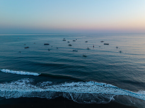 Aerial view of traditional fishing boats scattered across the calm ocean at dawn with gentle waves breaking on the dark sand beach in Buleleng, Bali, Indonesia.