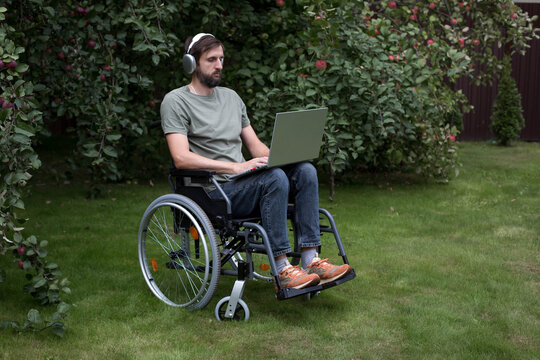 Bearded man in wheelchair uses laptop with headphones outdoors among apple trees, illustrating accessible work, remote employment, digital lifestyle, independence, inclusion, productivity, copyspace