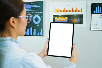 Businesswoman holding tablet with blank white screen mockup in front of colorful business analytics charts and graphs on office wall, ideal for technology and data presentation concepts © Stella 16