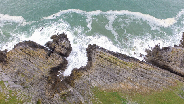Aerial view of the rugged limestone cliffs at Three Cliffs Bay with white sea foam crashing against the dark rock formations and turquoise water in the United Kingdom.