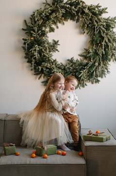 Happy sister and little brother hugging and laughing on couch with Christmas gifts and tangerines under a wreath made of christmas tree branches and garland lights in a decorated home livingroom.