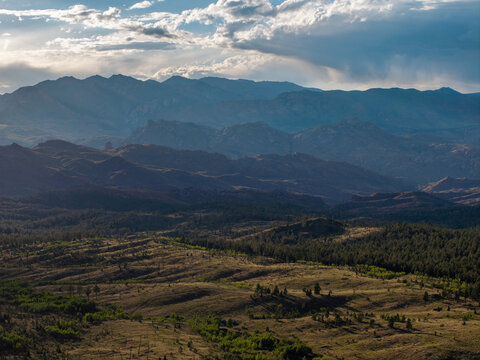 Aerial view of the Rocky Mountains featuring rolling hills, dense pine forests, and dramatic clouds under a bright sky in Colorado, United States.