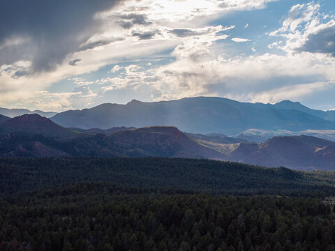 Aerial view of the Rocky Mountains featuring dense evergreen forests, rolling foothills, and dramatic storm clouds illuminated by soft sunlight in Colorado, United States.
