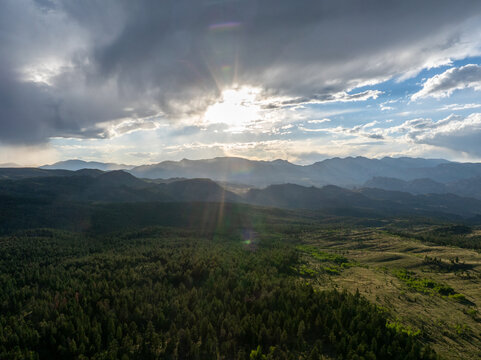 Aerial view of the Rocky Mountains in Colorado, United States, featuring dense pine forests, rolling hills, and dramatic sun rays breaking through storm clouds.