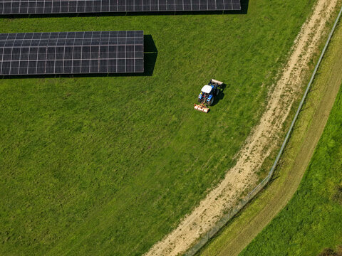 The Basin, Victoria, Australia - 18 September 2026: Aerial view of a blue tractor working in a green field next to rows of solar panels and a dirt path.