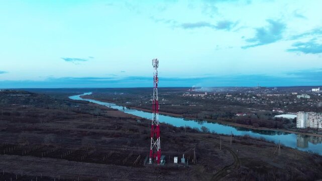 Red-and-white cellular communications tower in a drone arc over riverside fields. Telecommunications infrastructure, mobile broadband and community connectivity
