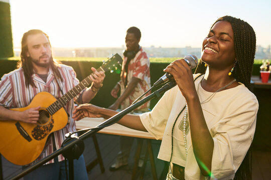 Young adult African American woman singing into microphone with multiethnic band playing acoustic guitar outdoors. Useful for music festival promo, entertainment advertising