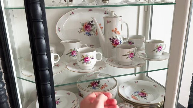 A woman's hand opens the door of a vintage sideboard containing antique porcelain tableware, picks up a coffee mug from the shelf