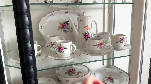 A woman's hand places a coffee mug on a shelf and closes the door of a vintage sideboard containing antique porcelain tableware