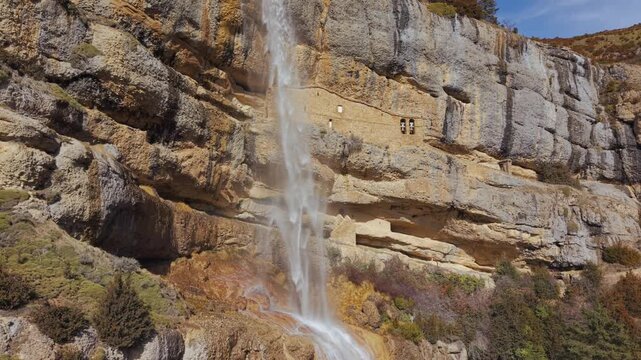 Aerial semi-orbit revealing a waterfall and the Iglesia de Las Cuevas and San Cornelio hermitages embedded in the cliff at Yebra de Basa, Spain.