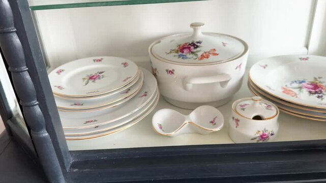 Antique porcelain tableware: a set of plates, a soup tureen, serving dishes with floral patterns on a shelf in a vintage sideboard