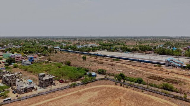 Aerial drone view showing a long train traveling along parallel railway tracks through dry countryside. Open land trees and distant buildings surround the railway corridor in Chennai India