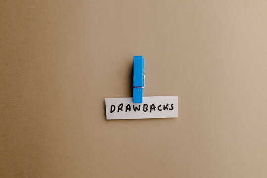 A small blue wooden clothespin hold a white paper strip with handwritten text drawbacks on a neutral brown background in minimalist studio style