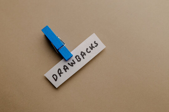 A conceptual photograph featuring a paper slip with the word drawbacks held by a blue clothespin on a plain neutral background surface