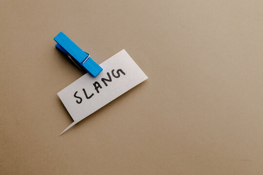 A handwritten word slang on a paper speech bubble attached by a bright blue wooden clothespin on a plain neutral background overhead shot