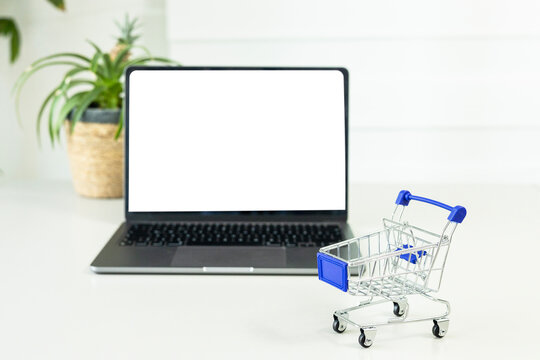 Blank white screen computer and mini shopping cart on table. Online shopping concept. Laptop mock up screen view.