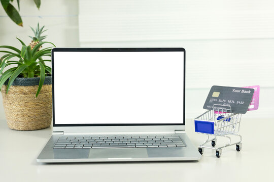Blank white screen computer, credit card and mini shopping cart on table. Online shopping concept. Laptop mock up screen view.