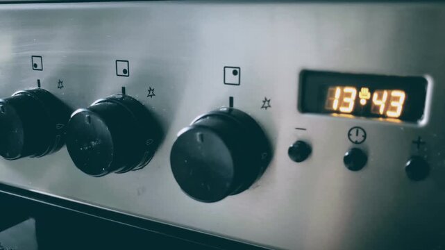 Close up of stainless steel oven control panel with knobs and digital clock. Candid camera pan right shot of modern kitchen appliance in sleek light.