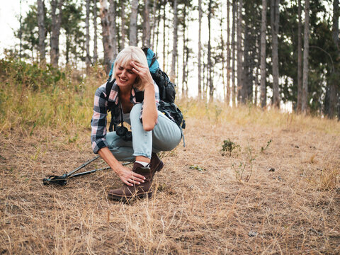 Woman hiker suffering from ankle pain during mountain hike, stopping to rest on forest footpath while dealing with injury, fatigue and difficulty walking
