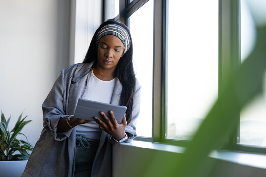 African woman leaning on window ledge in office wearing blazer using gray tablet with plant nearby