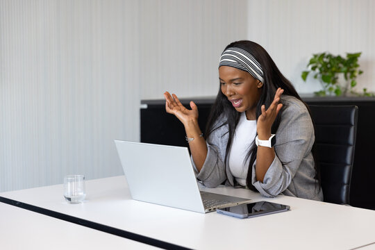 African woman wearing blazer, striped headband raising hands at office table with laptop