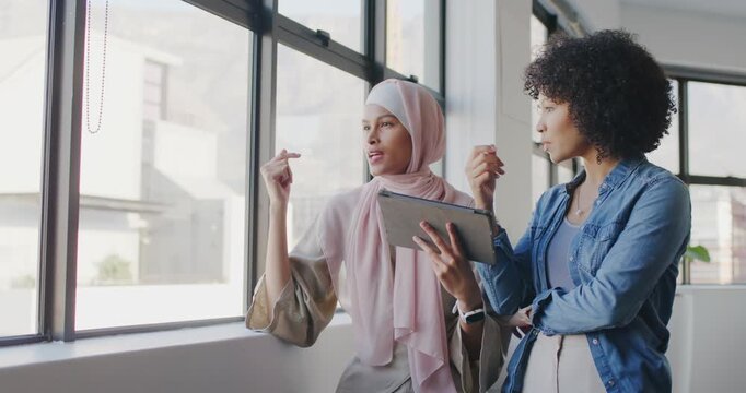Diverse female coworkers in hijab, denim pointing to view, sharing tablet at office finalizing plan