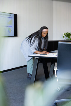 African American woman in gray blazer marking papers with marker at meeting table viewing screen